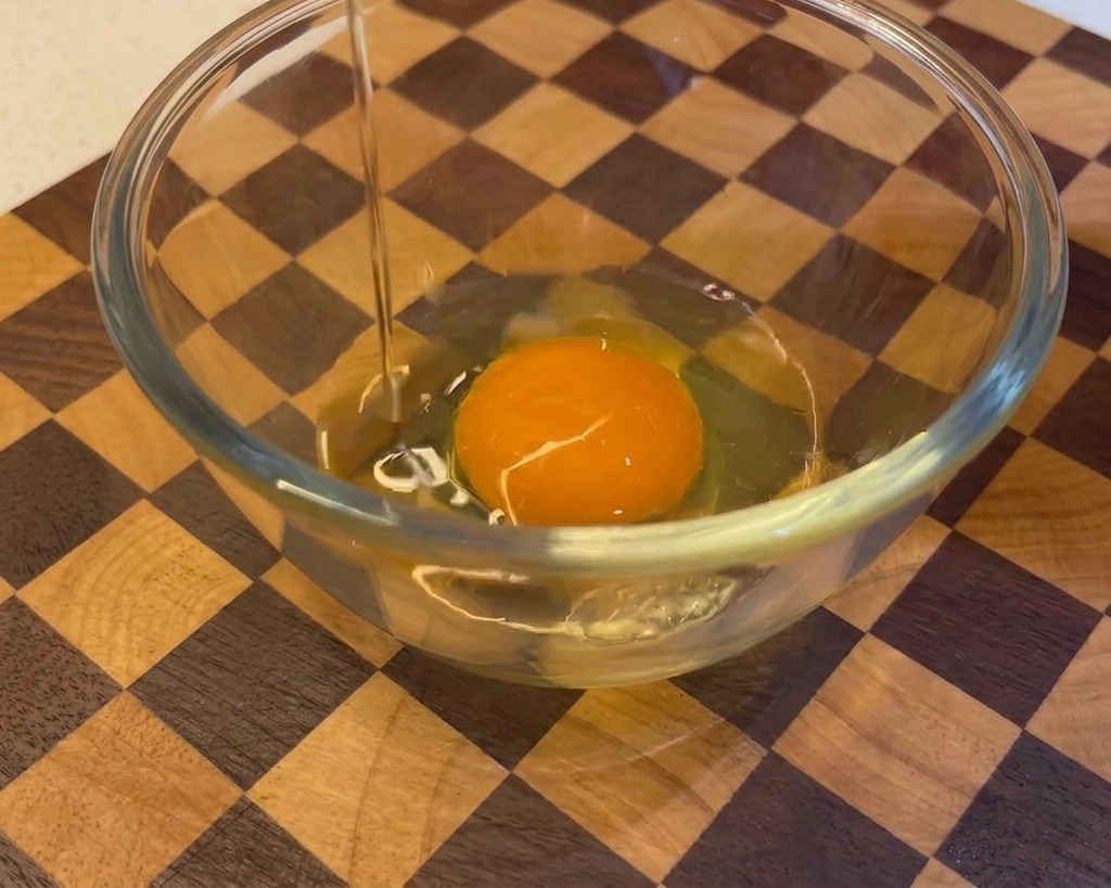 Un uovo crudo che viene rotto e versato in una ciotola di vetro che contiene già un tuorlo, su uno sfondo di legno.