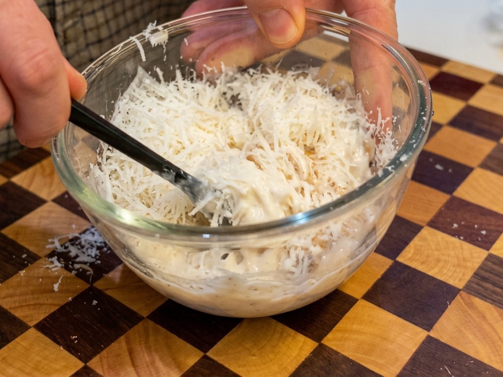 Un composto denso e cremoso di uova e formaggio giallo pastello mescolato con bacchette nere in una ciotola trasparente.