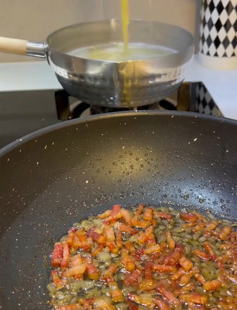 Una mano usa pinze di metallo per sollevare gli spaghetti gialli da una pentola di acqua bollente e trasferirli in una padella con guanciale e cipolle.