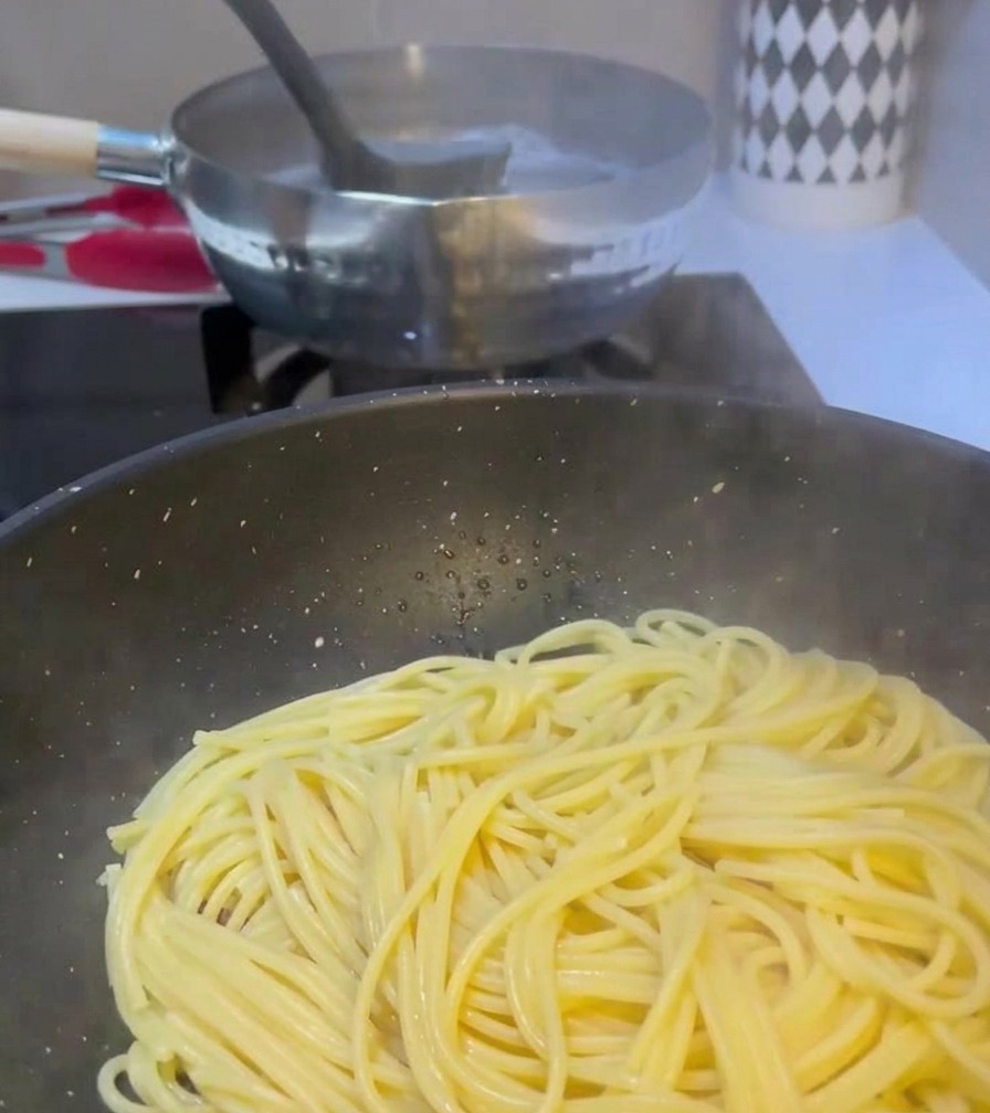 Un mestolo che trasferisce lacqua opaca da una pentola bollente di pasta a una padella che contiene già spaghetti e carne cotta.
