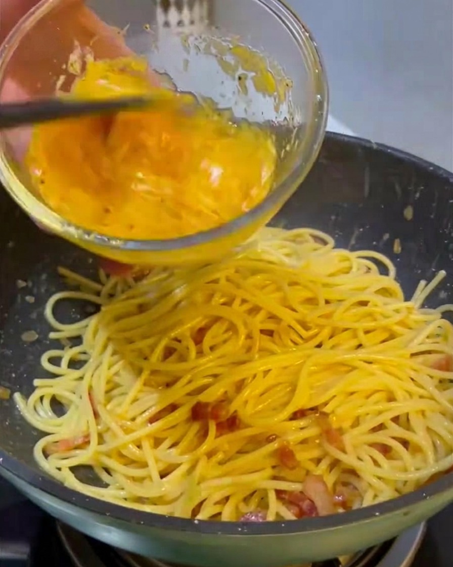 Versare un composto giallo vivo di uova e formaggio da una ciotola di vetro su una padella di spaghetti caldi e guanciale.