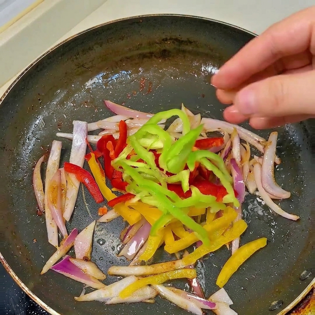 Una mano aggiunge striscioline di peperone rosso, giallo e verde in una padella nera con le cipolle già appassite.