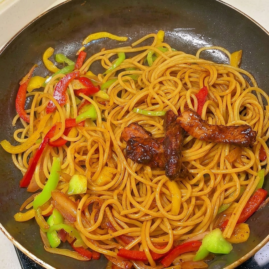 Straccetti di maiale saltati adagiati sopra gli spaghetti al pepe nero e peperoni in una grande padella.