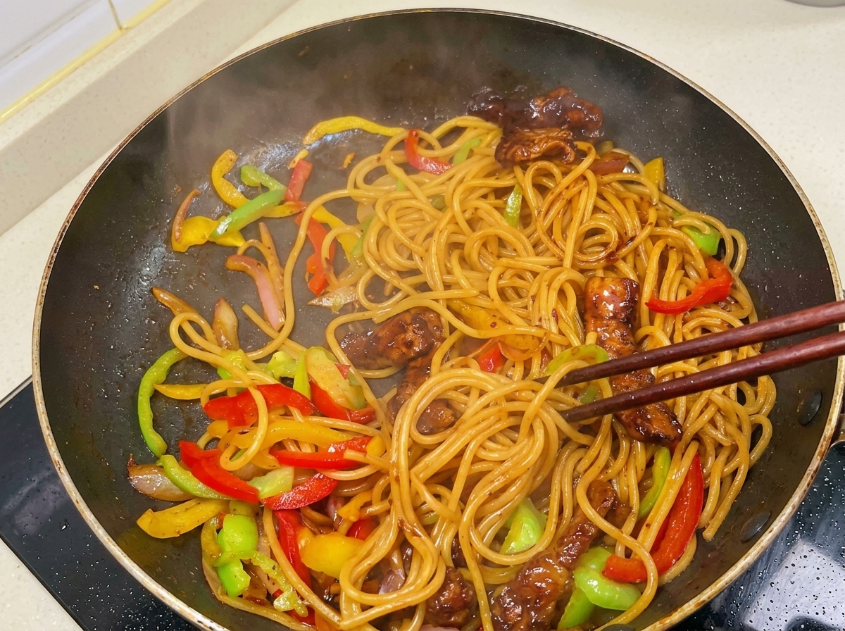Bacchette che saltano un mix fumante di spaghetti, straccetti di maiale rosolati e peperoni a fette, il tutto avvolto da una salsa scura e lucida.