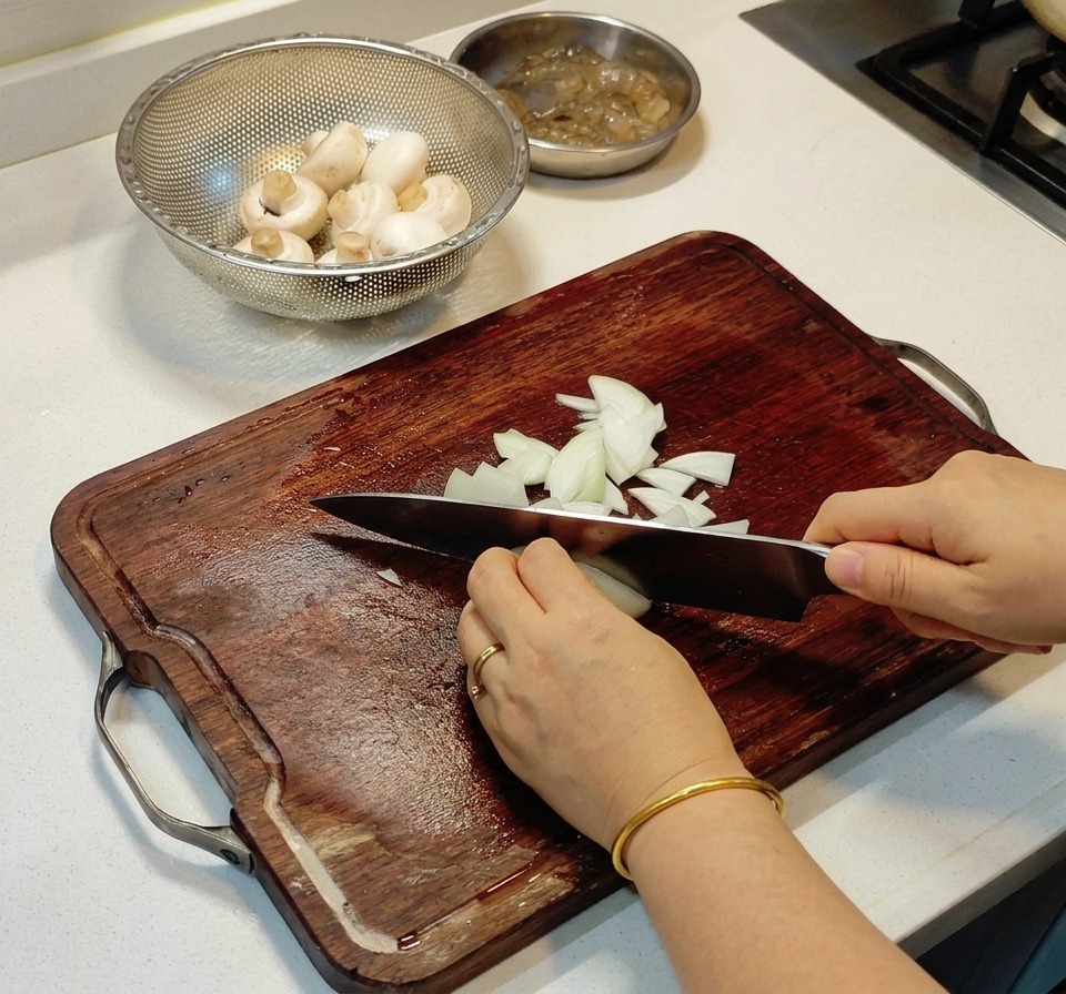 Mani che usano un coltello da chef per tritare finemente una cipolla bianca su un tagliere di legno.