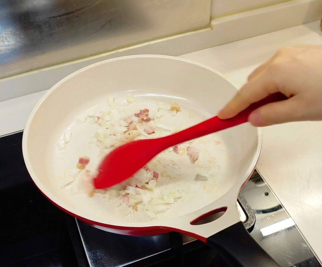 Una spatola rossa che mescola cipolla tritata e pezzetti di bacon in una padella bianca con rivestimento in ceramica.