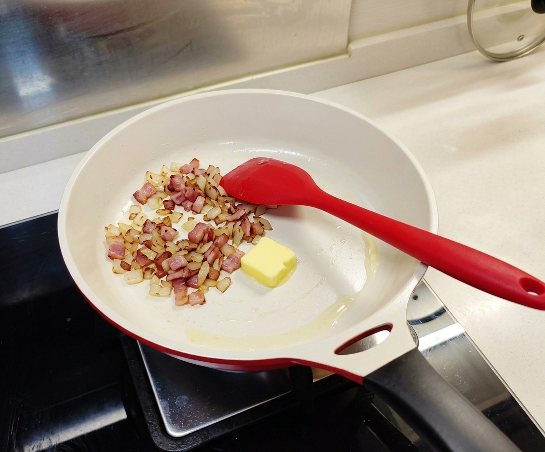 Un cubetto di burro giallo in una padella bianca insieme alla cipolla e al bacon già cotti.