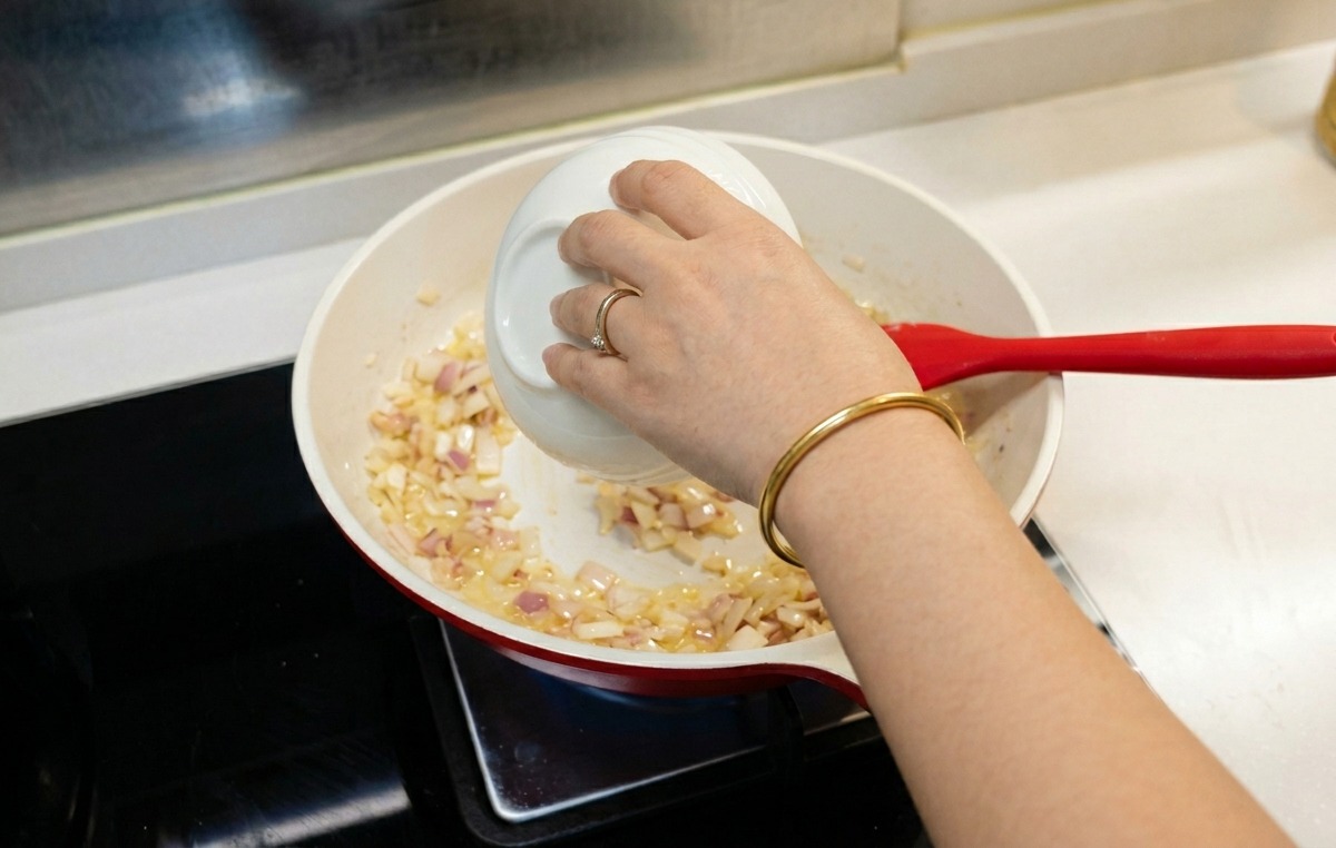 Una persona che versa dellacqua da una ciotolina in una padella con cipolle e farina cotte.