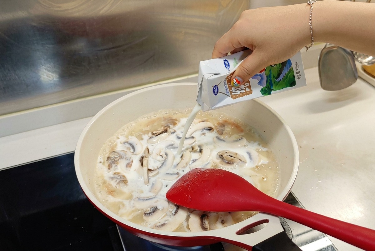 Latte versato da un cartone bianco e azzurro in una padella in cui sobbollono gamberi e funghi.