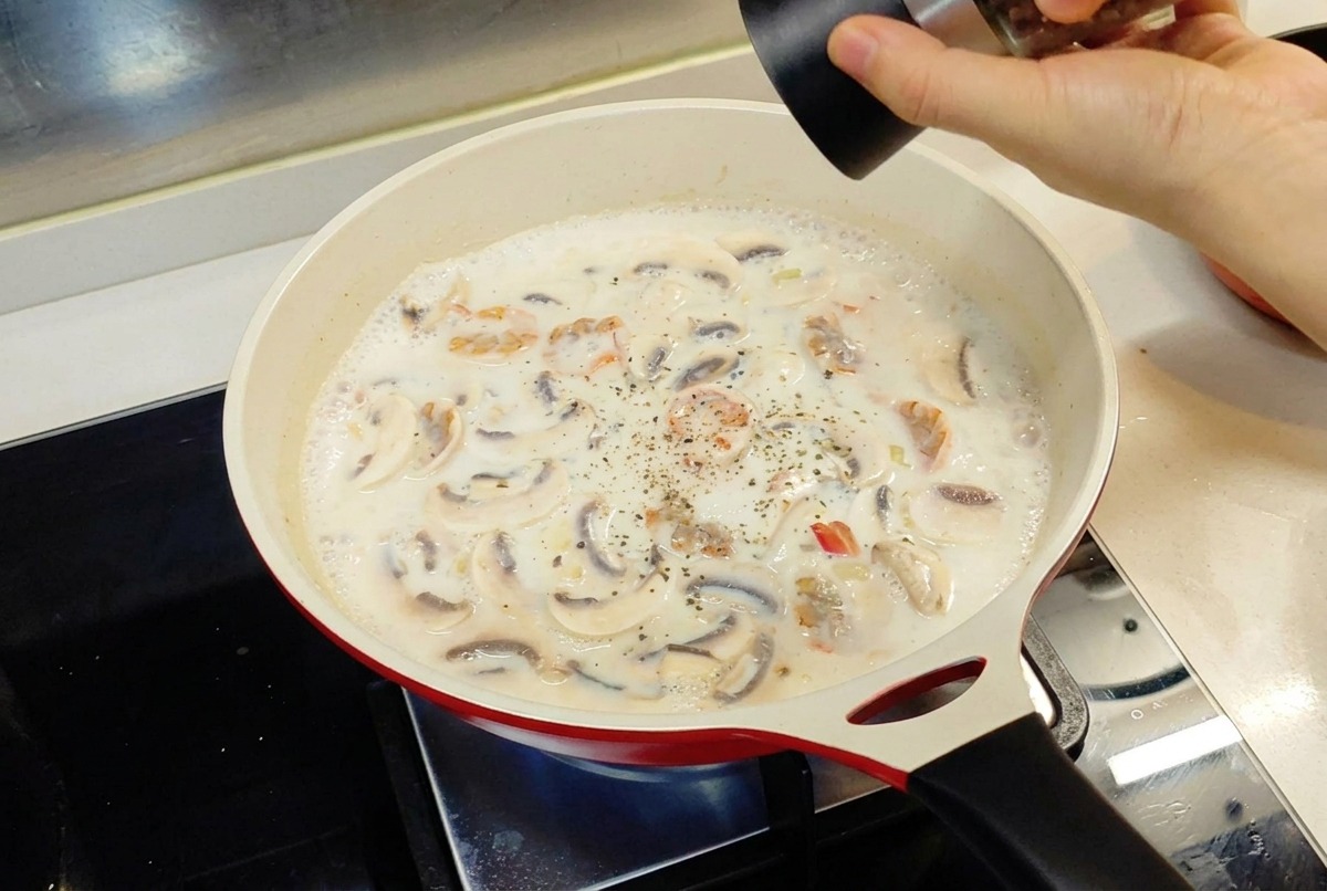 Una mano che usa un macinapepe per aggiungere pepe nero a una salsa bianca che sobbolle in una padella rossa.