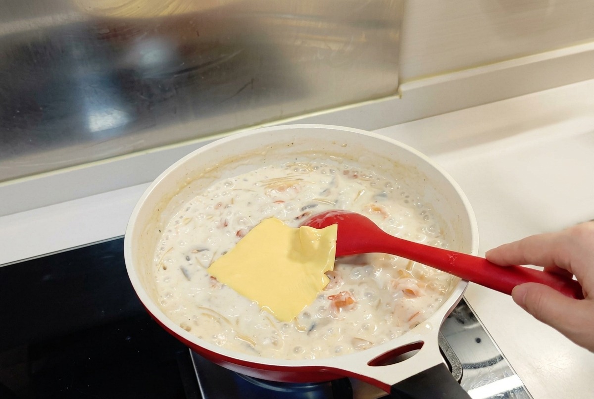 Una fetta quadrata di formaggio fuso poggiata sopra la pasta alla salsa bianca in una padella con un cucchiaio rosso.