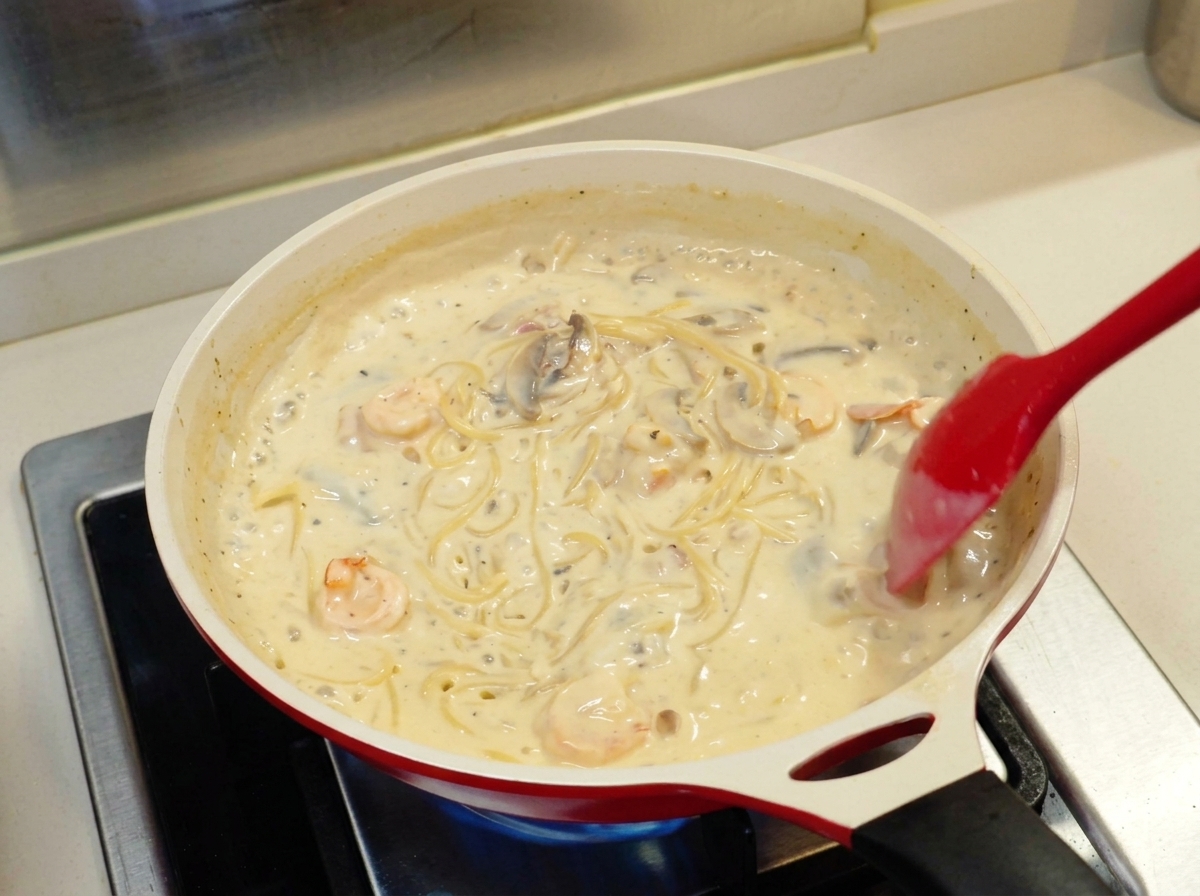 Una spatola rossa che gira gli spaghetti in una densa salsa bianca con gamberi e funghi in una padella di ceramica.