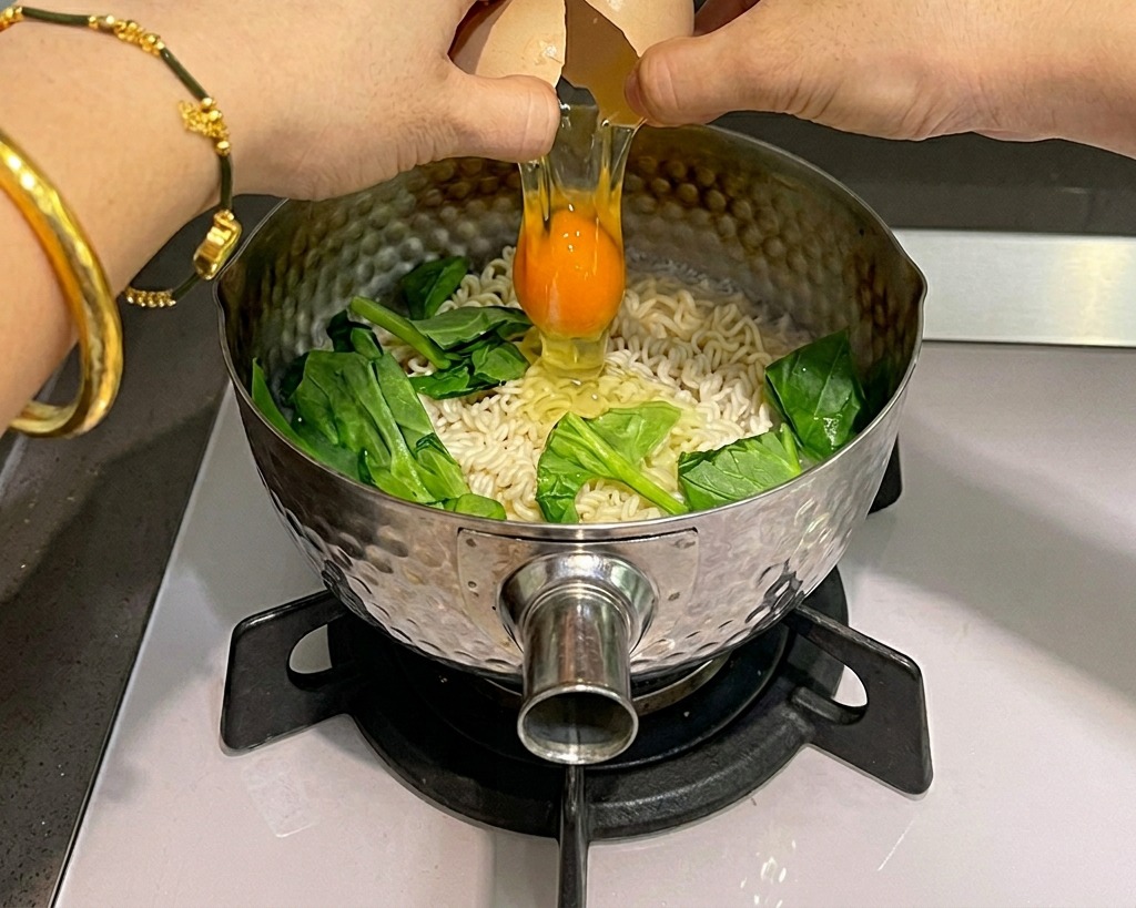 Due mani che rompono un uovo crudo sopra una pentola di ramen istantaneo e spinaci verdi in cottura.