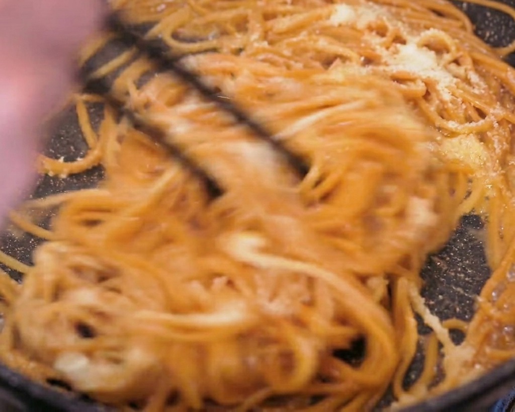 Spaghetti integrali saltati energicamente in padella con delle bacchette nere, mostrando il formaggio e il burro che si sciolgono in una crema.
