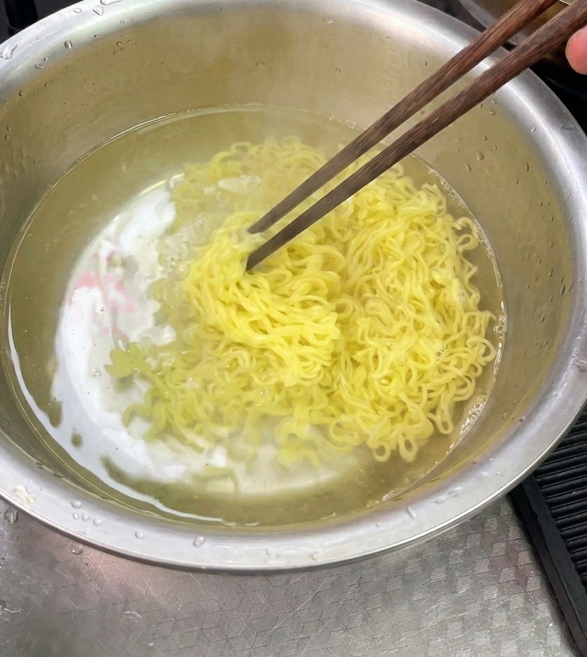Noodles gialli cotti sciacquati e mescolati con le bacchette in una ciotola di acciaio inossidabile piena dacqua fredda.