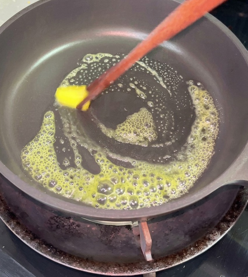Un cubetto di burro giallo che si scioglie e sfrigola in una padella antiaderente nera, mosso con bacchette di legno.