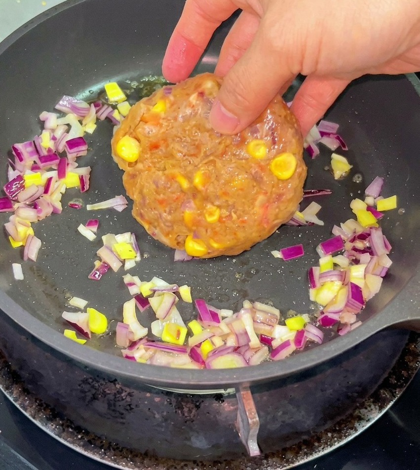 Una mano che inserisce un tortino di carne di manzo cruda e mais in una padella con cipolle rosse tritate e burro fuso.