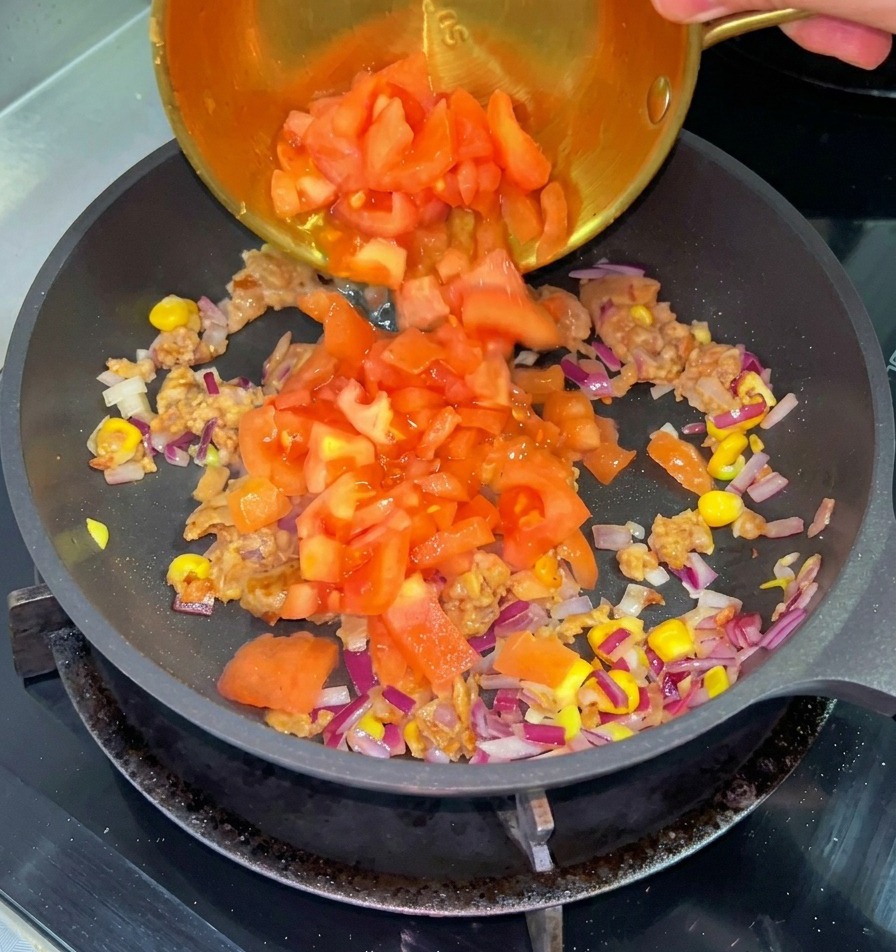 Pomodori rossi a cubetti versati da una piccola ciotola di ottone in una padella con manzo cotto, mais e cipolle rosse.