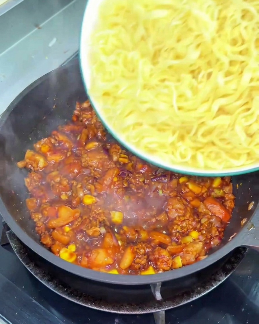 Una ciotola di noodles gialli ondulati versati in una padella piena di un sugo denso di pomodoro e manzo.