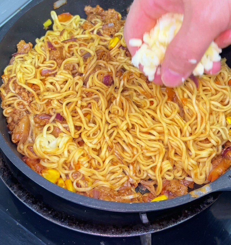 Un primo piano di una mano che cosparge di formaggio bianco grattugiato una padella di noodles saltati con carne macinata, cipolle e mais.