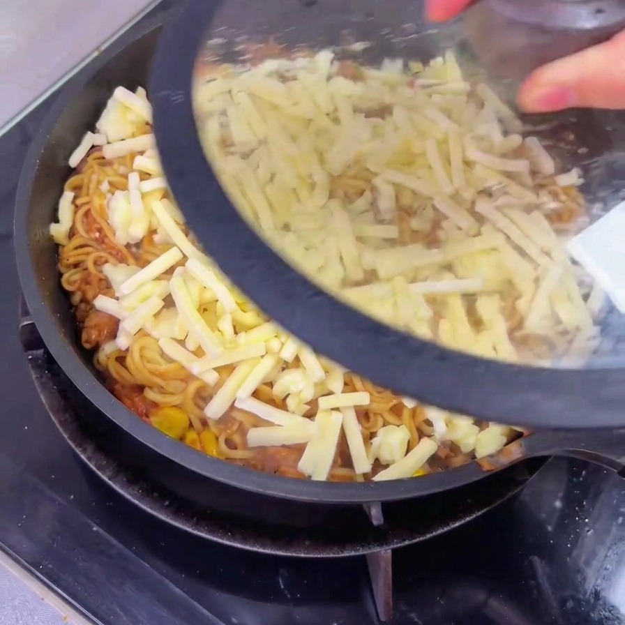 Una persona che posiziona un coperchio di vetro trasparente su una grande padella contenente noodles al manzo ricoperti da uno strato di formaggio grattugiato non ancora sciolto.