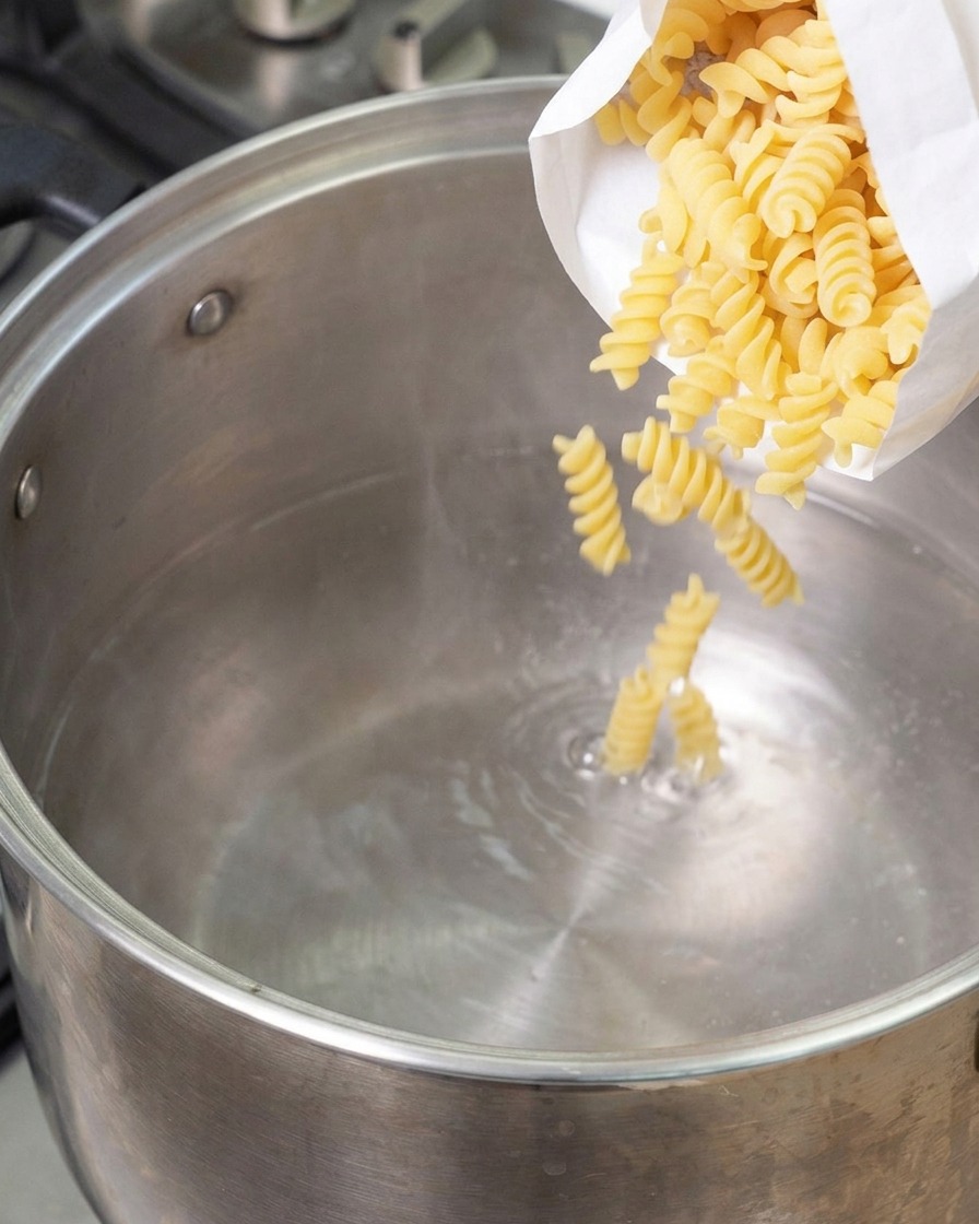 Fusilli secchi versati da un sacchetto bianco in una grande pentola di acciaio con acqua bollente.