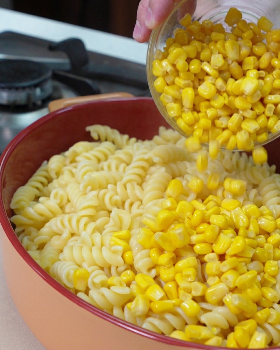 Una ciotola di vetro da cui vengono versati chicchi di mais dolce giallo brillante sopra i fusilli cotti in una pirofila rotonda.