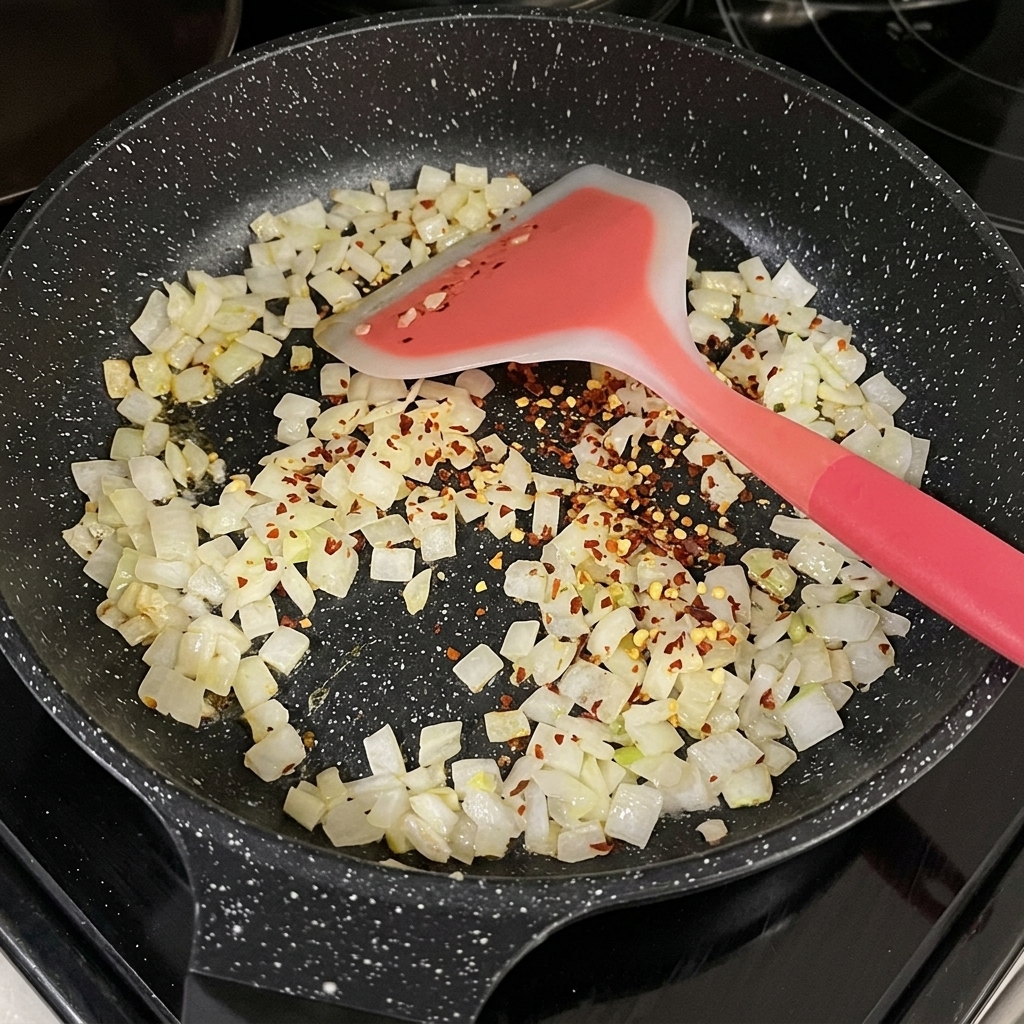 Peperoncino rosso secco a scaglie sparso sulla cipolla a cubetti appassita in una padella nera.