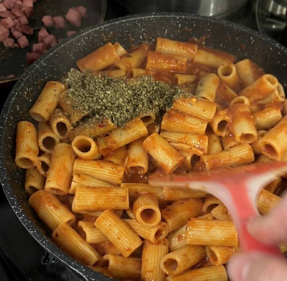 Una generosa manciata di basilico essiccato a scaglie aggiunta sopra i rigatoni al pomodoro in una padella nera.