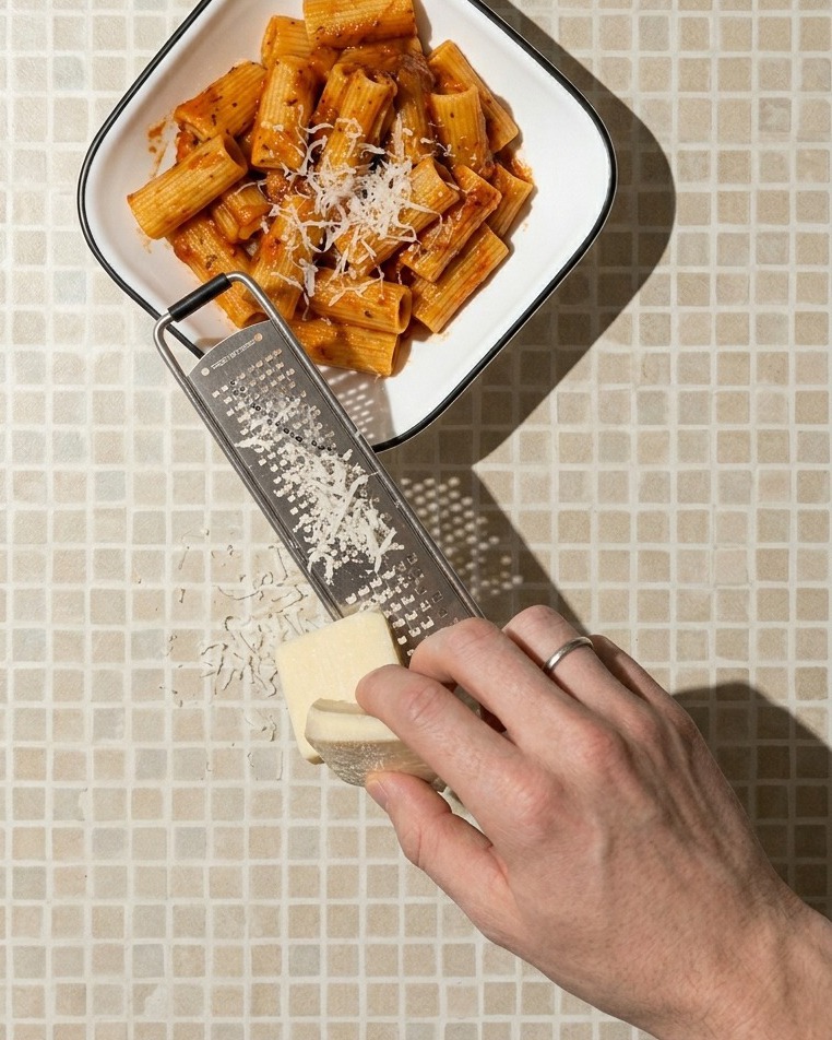 Primo piano di una mano che grattugia un pezzo di formaggio chiaro sopra una ciotola quadrata bianca piena di rigatoni al pomodoro.