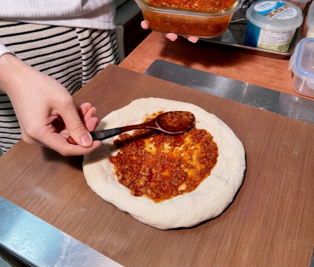Un cucchiaio di legno stende un denso ragù di carne rossastro con un movimento circolare sulla base cruda della pizza.