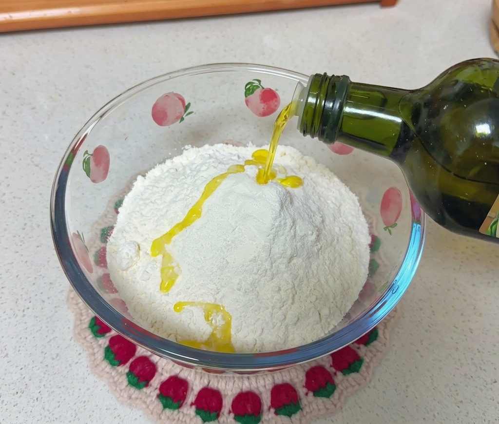 Olio doliva versato da una bottiglia di vetro verde scuro su una montagna di farina bianca in una ciotola di vetro decorata.