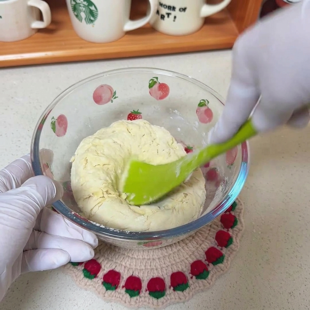 Una spatola verde che mescola un impasto grezzo di farina, acqua e olio in una ciotola di vetro trasparente con motivi di frutta.