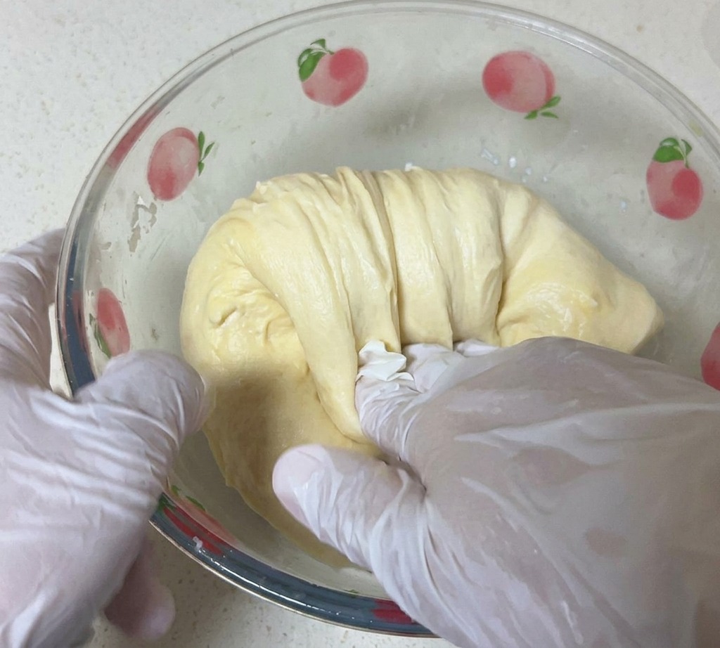 Una persona con guanti bianchi solleva e allunga un impasto liscio e giallo chiaro allinterno di una ciotola di vetro.