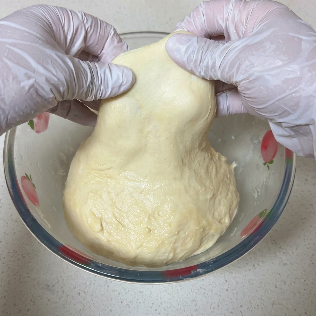 Due mani con guanti bianchi tendono delicatamente un lembo di impasto per pizza mostrando una membrana sottile e trasparente in controluce.