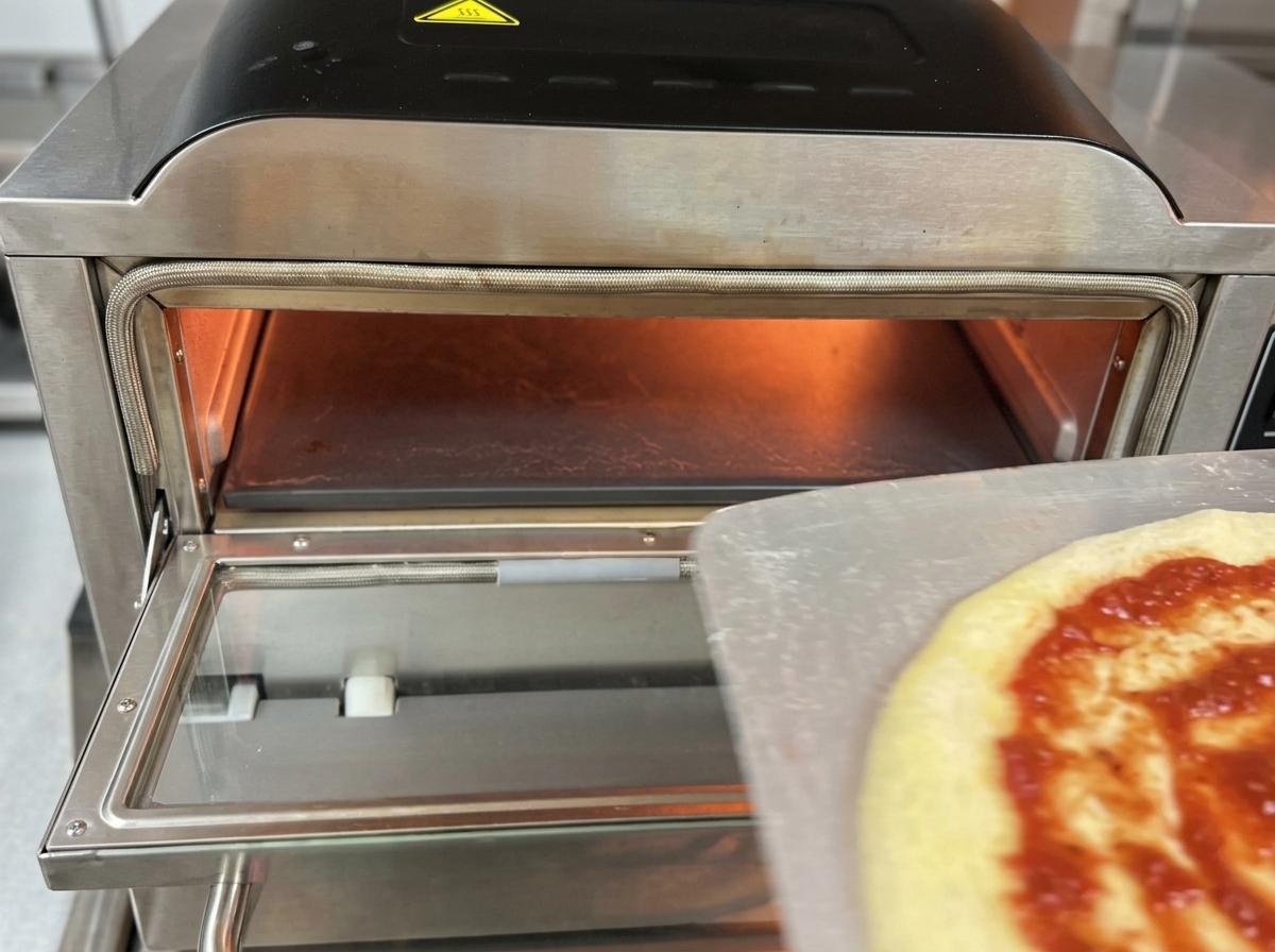 Una pizza cruda condita con pomodoro e mozzarella mentre viene trasferita da una pala di metallo allinterno di un forno elettrico professionale per pizza.