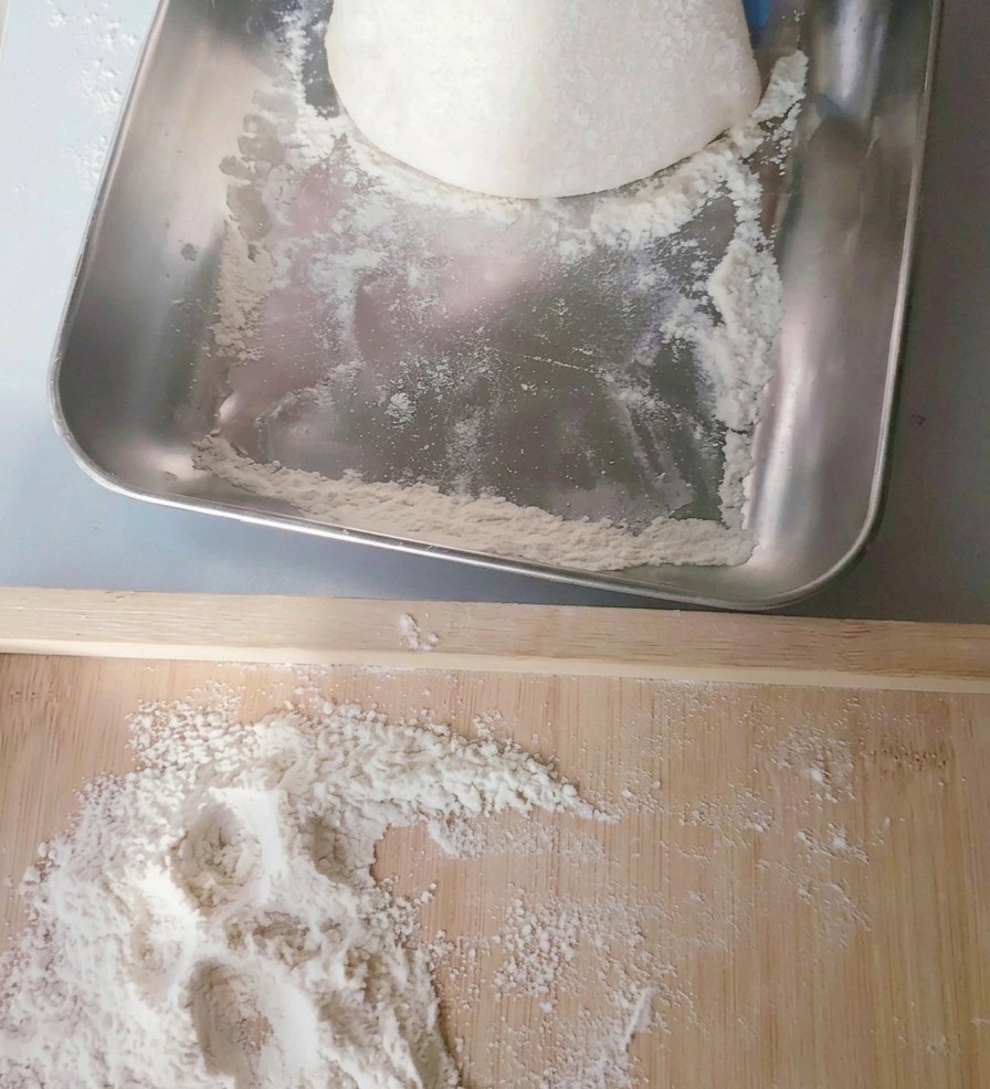Un panetto rotondo di impasto per pizza prelevato da una cassetta in acciaio e adagiato su una spianatoia di legno infarinata.