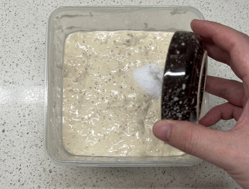Una mano versa il sale da una piccola ciotola in un contenitore di plastica con il poolish bolloso.
