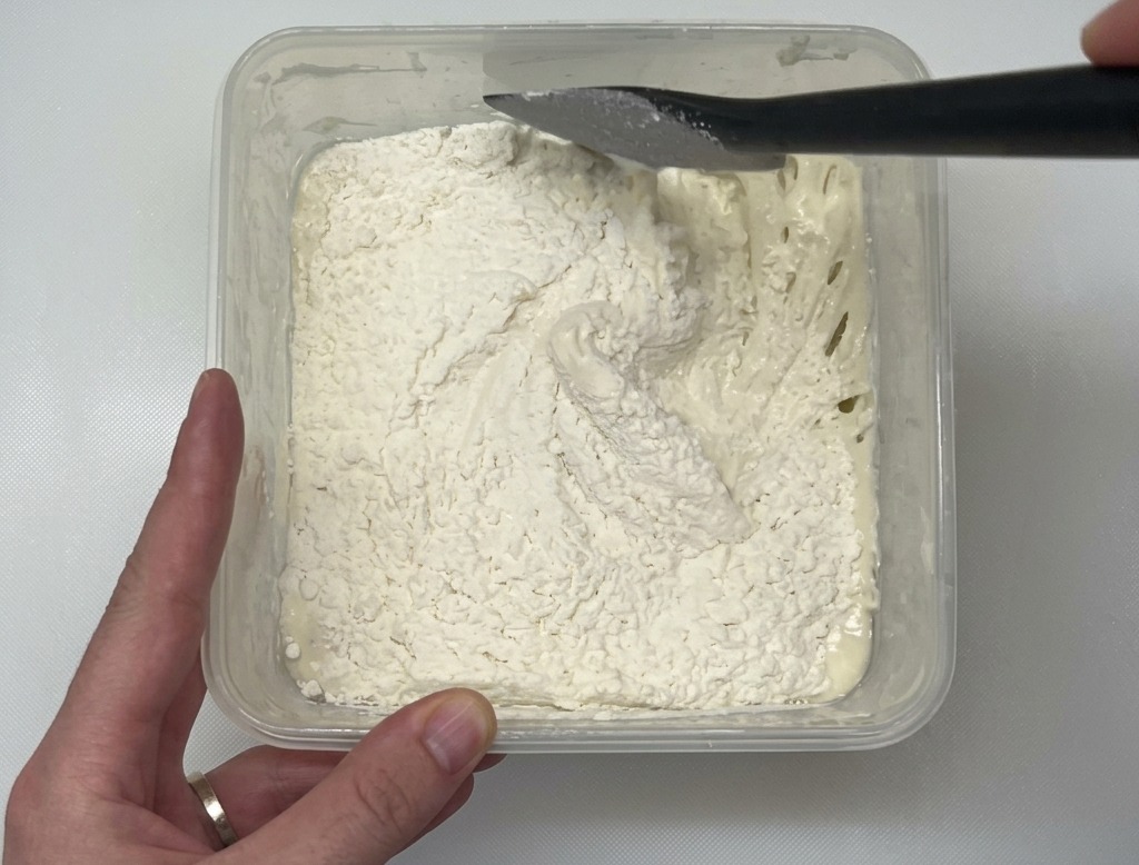 Una spatola nera mescola la farina bianca in un impasto umido dentro un contenitore trasparente.