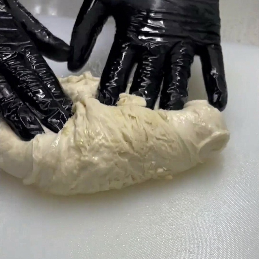 Una persona con guanti neri stende e piega un pezzo di impasto chiaro e umido su una superficie bianca.