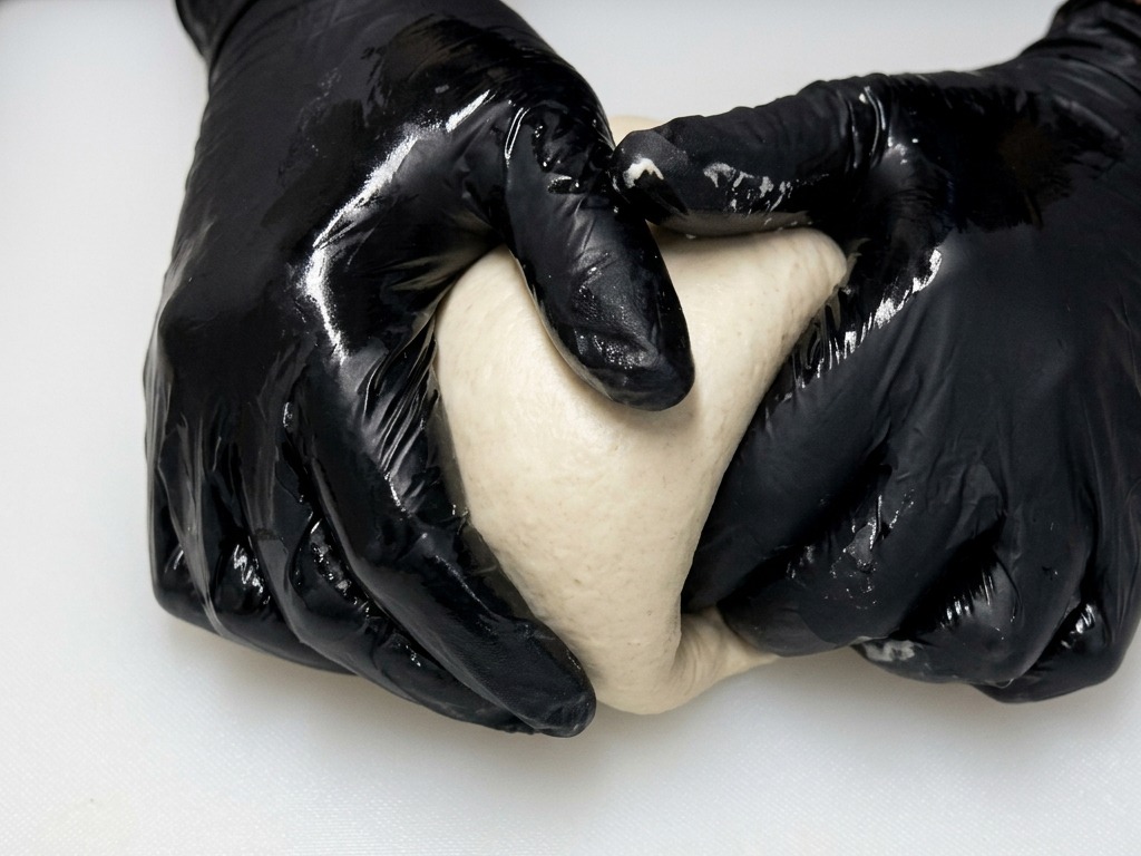 Mani con guanti neri formano una pallina di impasto liscia e rotonda facendola scivolare su un piano bianco.
