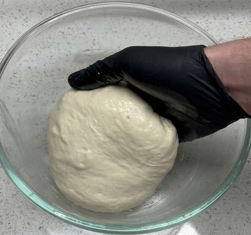 Una mano con un guanto nero mette un panetto liscio in una grande ciotola di vetro sul bancone.