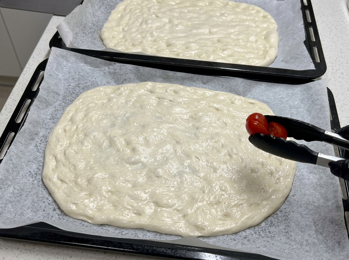 Usando pinze da cucina nere, un pomodorino tagliato a metà viene posizionato sulla base di pizza stesa.