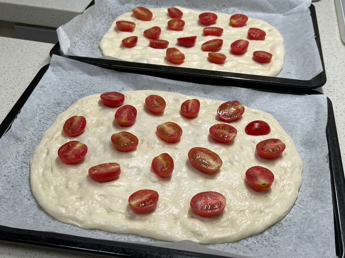 Due teglie mostrano limpasto della pizza steso e condito con numerosi pomodorini tagliati a metà, pronto per il condimento.