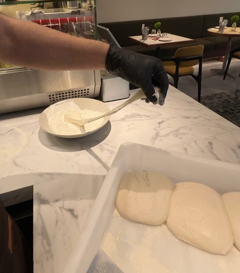 Una mano con guanto che solleva un panetto elastico di impasto per pizza da una ciotola piena di farina bianca.