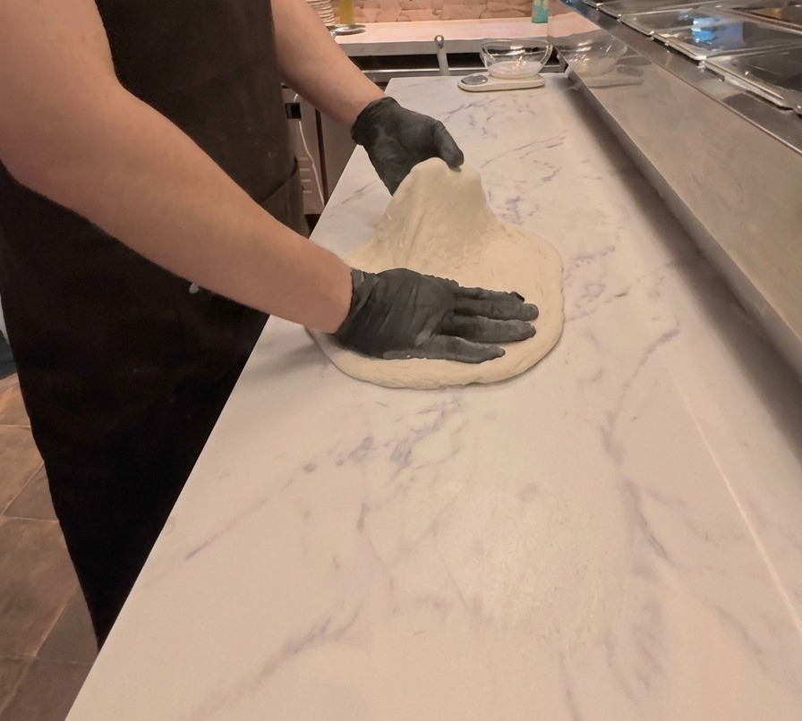 Un cuoco che usa entrambe le mani per stendere e appiattire limpasto della pizza su un bancone di marmo bianco.