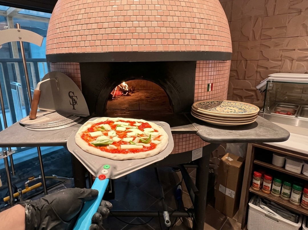 Una pizza cruda condita con pomodoro e mozzarella adagiata su una pala metallica allingresso di un forno a legna rivestito di piastrelle rosa.