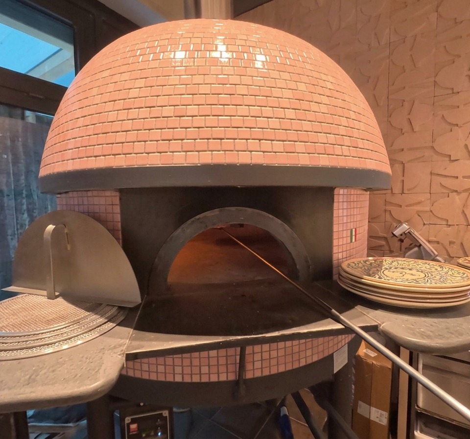 Lesterno di un forno a cupola per pizza con piastrelle rosa, con una pizza visibile in cottura allinterno vicino alle braci ardenti.