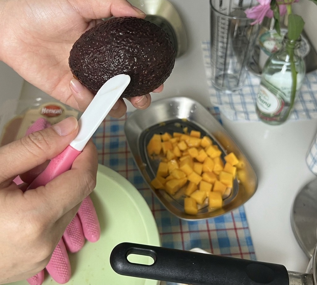 Mani che tagliano a cubetti una metà di avocado sbucciato sopra un vassoio con il mango a dadini.