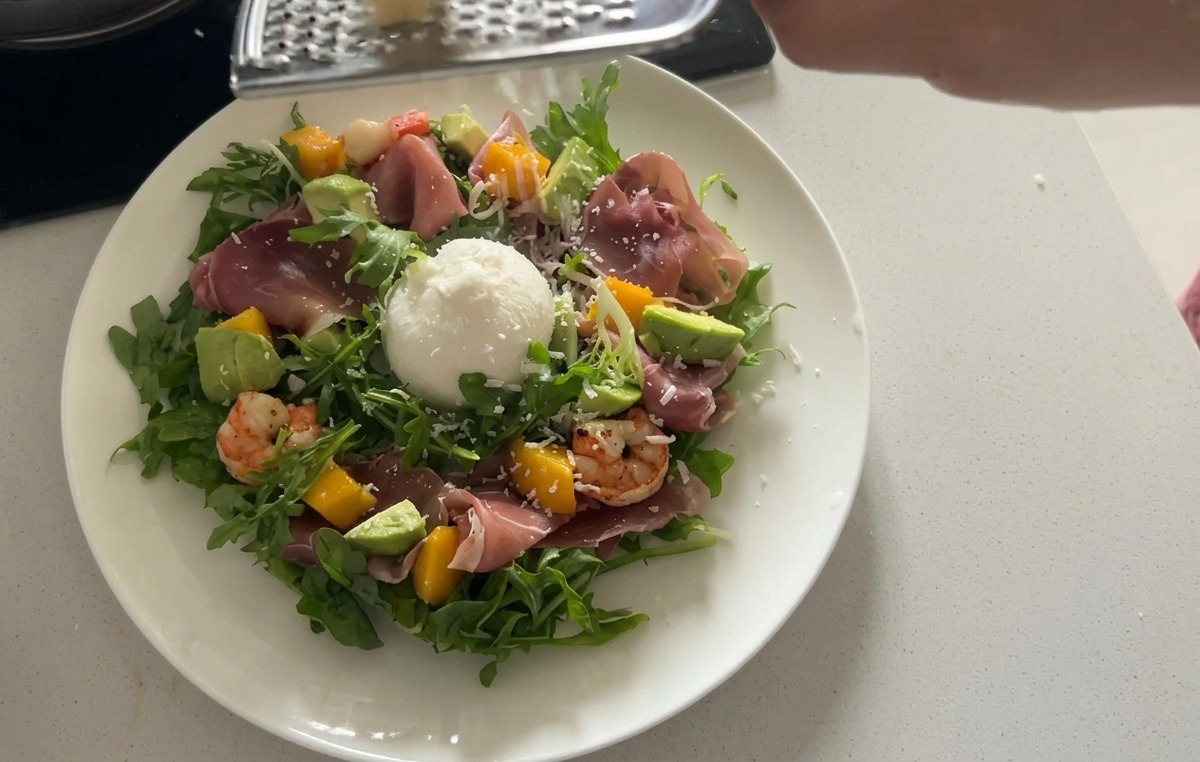 Insalata di rucola impiattata e arricchita con una grande burrata bianca e sottili fette di prosciutto crudo.